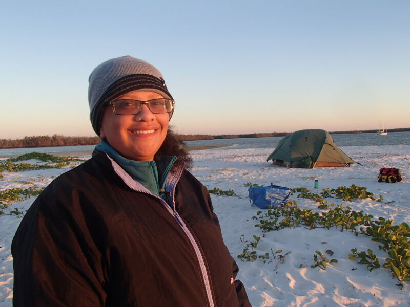 A woman wearing glasses and a beanie stands in a snowy landscape, smiling at the camera. Behind her, there's a tent pitched on the snow-covered ground, suggesting a camping scene. The sky is clear and bright, possibly during sunrise or sunset, casting a warm glow on the surroundings. The overall impression is one of enjoying the outdoors in a winter setting.